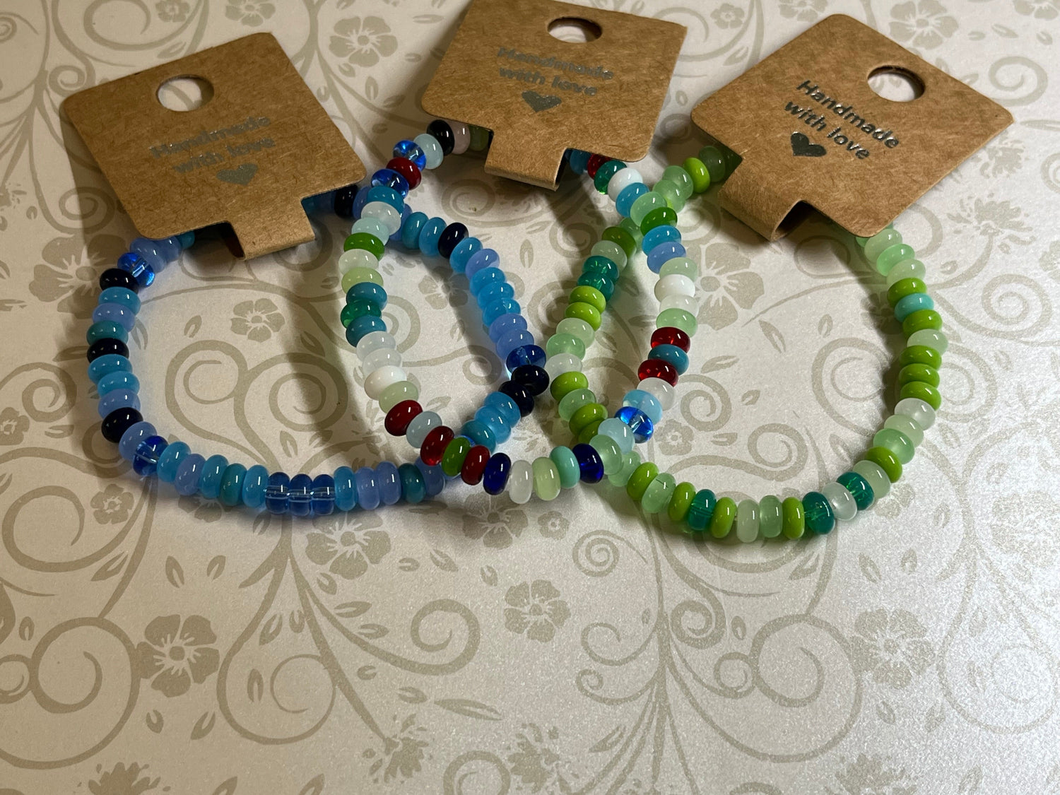 Three handmade abacus bead bracelets in blues and greens, tied onto kraft cards stamped Handmade with love